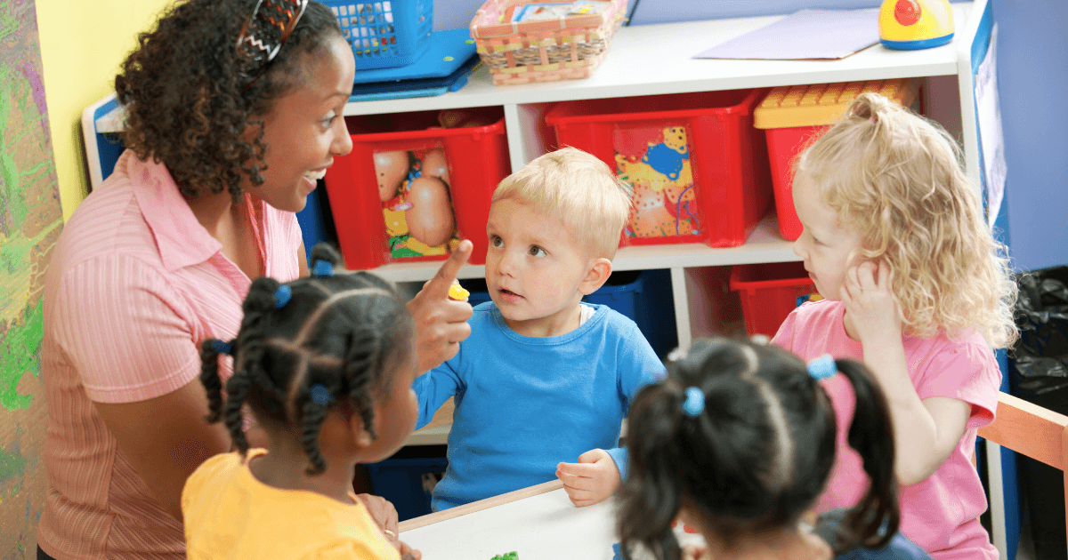 A teacher smiles and interacts with four young children seated around a table in a colorful classroom. Using the Ascend Method, the children are engaged, with toys and storage bins visible in the background.