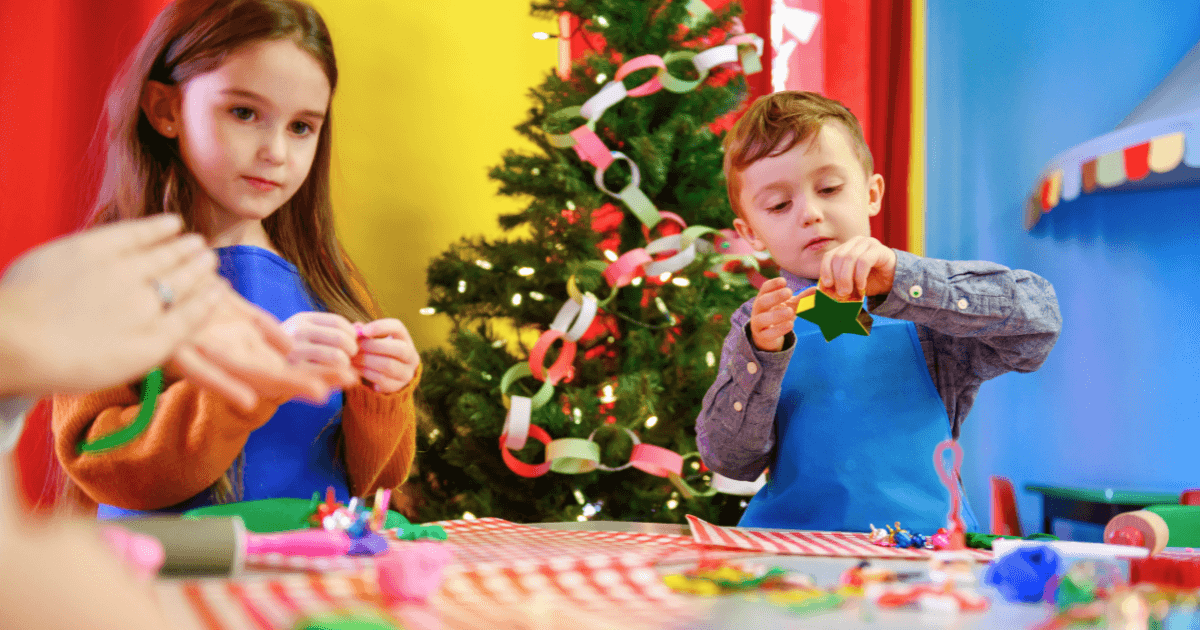Two children wearing blue aprons sit at a table making holiday crafts with colorful paper, celebrating December celebrations in front of a decorated Christmas tree and festive paper chain garland in a brightly colored room.
