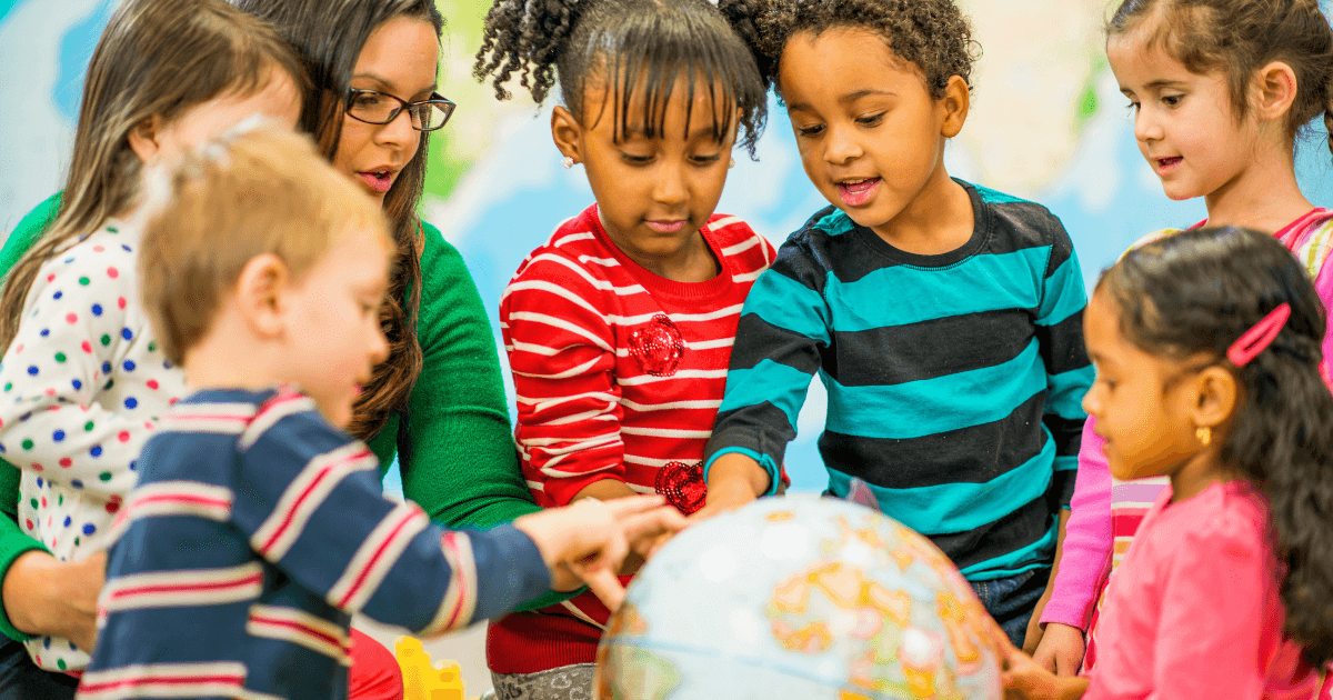A group of young children and a teacher gather around a globe, pointing and exploring together in celebration of Earth Day. The children appear curious and engaged, with colorful clothing and a blurred classroom background.