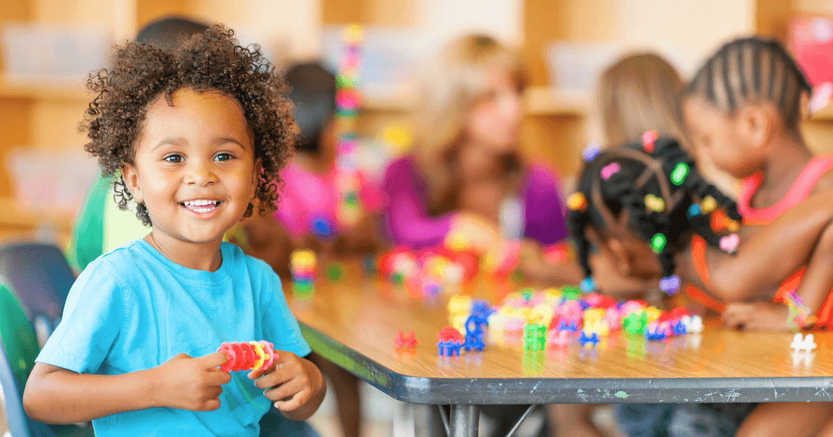 A young child smiles at the camera while holding colorful plastic toys, sitting at a table with other children who are playing and building in a classroom—discover joyful moments like this in Head Start programs near me.