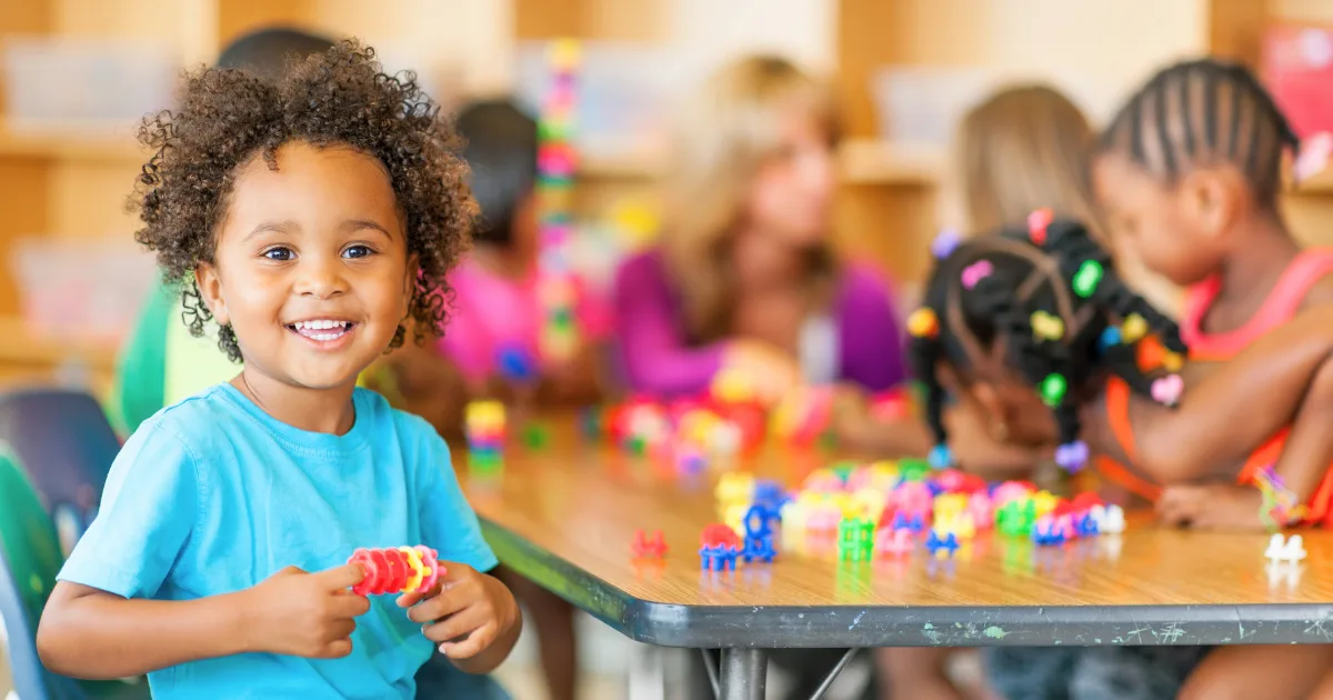 A young child smiles at the camera while holding colorful plastic toys, sitting at a table with other children who are playing and building in a classroom—discover joyful moments like this in Head Start programs near me.