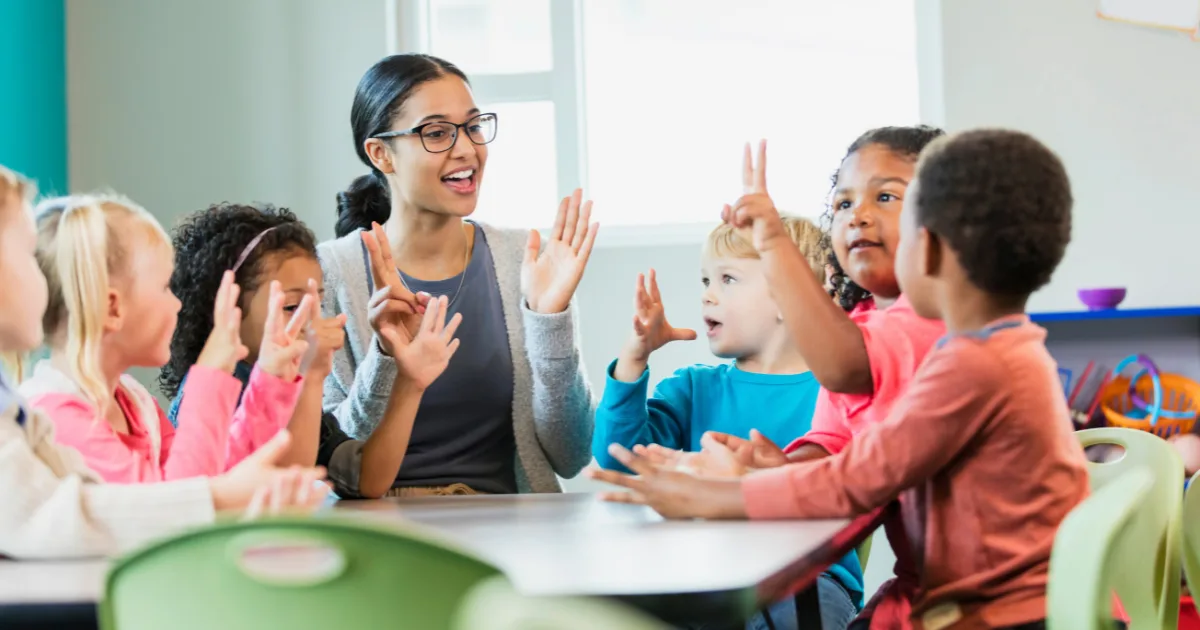 A teacher sits at a table with a group of young children, all raising their hands and smiling as they engage in a HighScope-inspired interactive classroom activity. The room is bright and cheerful.