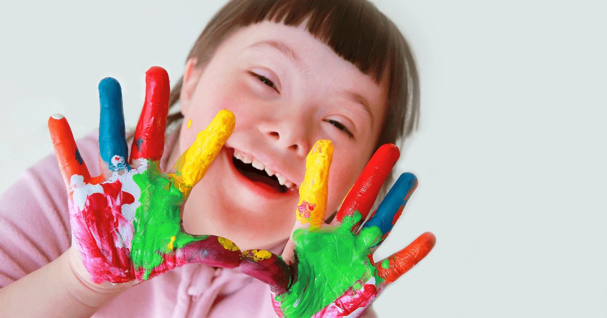 A smiling child with Down syndrome holds up both hands covered in brightly colored paint, showing joy and creativity in an inclusive preschool classroom. The plain, light background keeps the focus on the child’s happiness and artistic expression.