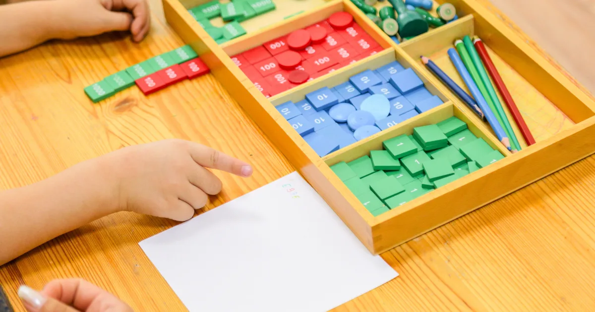 Two children’s hands are visible at a table with math manipulatives, colored sticks, and blank paper. The materials are organized in a wooden tray, perfect for Montessori at home hands-on math activities.