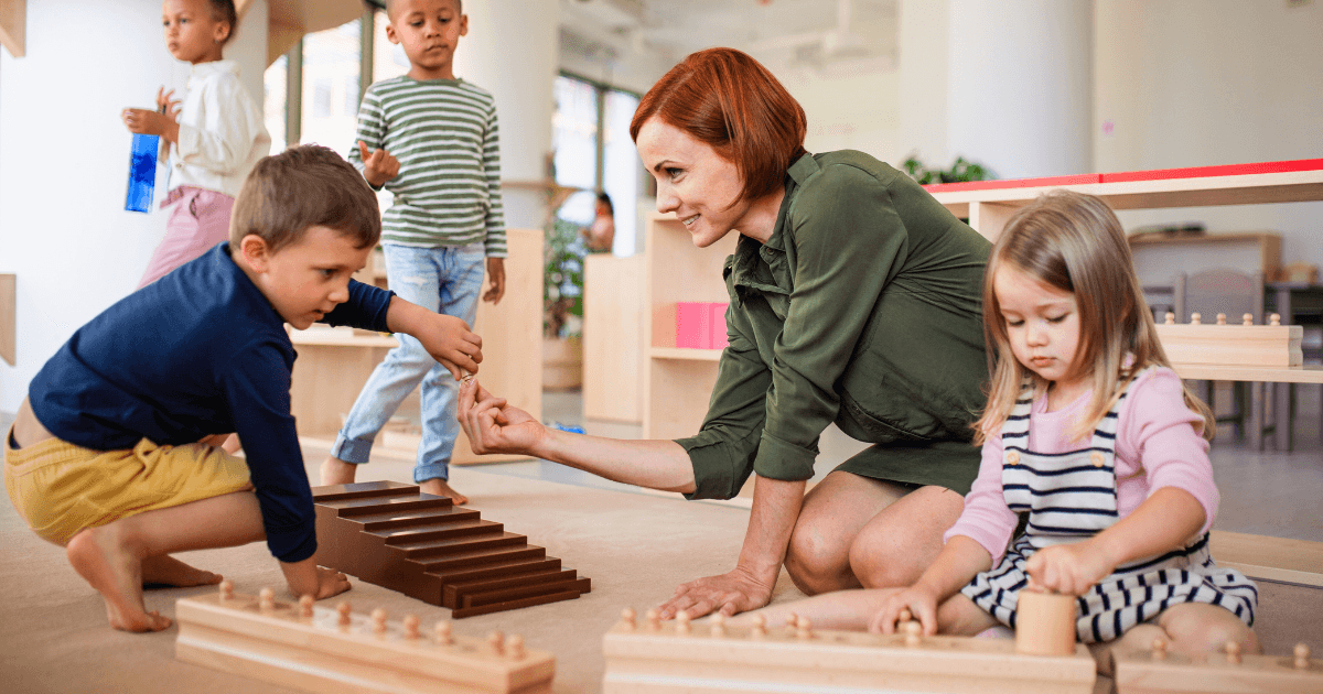 A teacher kneels on the floor with young children in a bright, inviting classroom, engaging with them as they play using Montessori method educational materials like wooden blocks and cylinders.