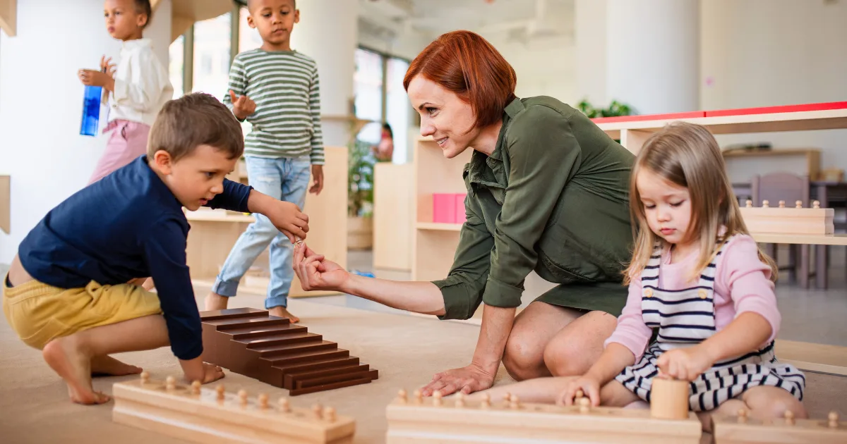 A teacher kneels on the floor with young children in a bright, inviting classroom, engaging with them as they play using Montessori method educational materials like wooden blocks and cylinders.