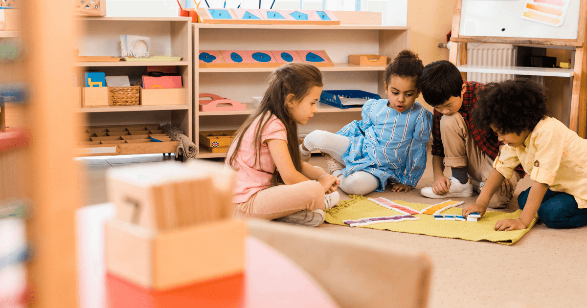 Four young children sit on the floor of a classroom in one of the Montessori schools near me, engaged in a learning activity with colorful cards and a yellow mat. Shelves with educational materials are in the background.