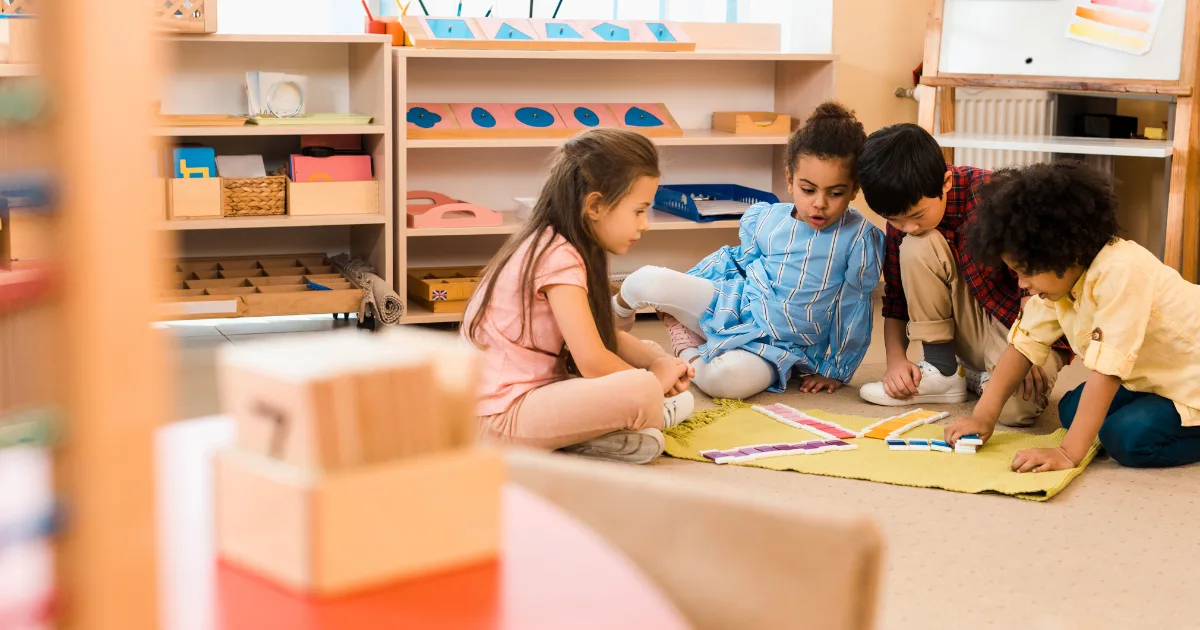 Four young children sit on the floor of a classroom in one of the Montessori schools near me, engaged in a learning activity with colorful cards and a yellow mat. Shelves with educational materials are in the background.