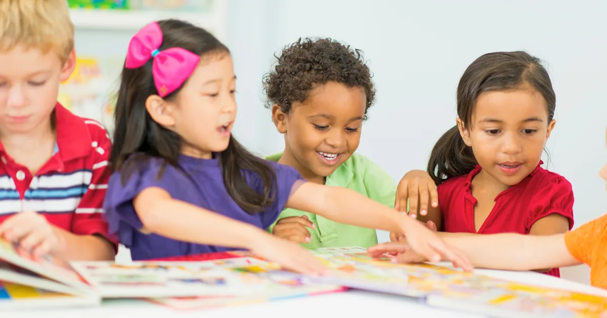 Four young children sit together at a table, smiling and looking at colorful picture books. One girl excitedly points at a page while the others watch and engage with the Pre-K curriculum materials.