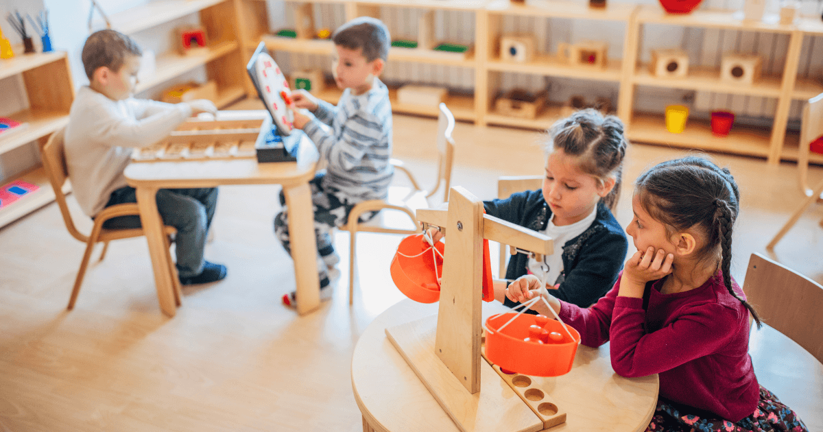 Four young children sit at small tables in a Reggio Emilia preschool classroom, engaging with educational toys. Two girls use a balance scale, while two boys work with puzzles and blocks in a bright, organized setting.