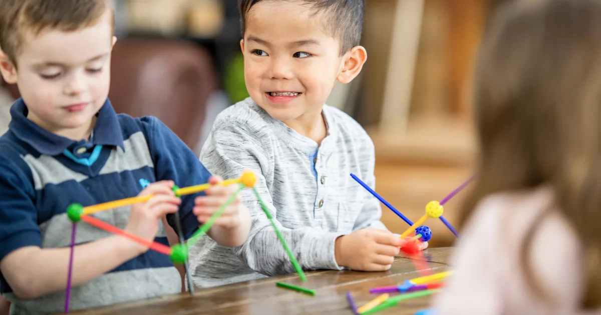 Three young children sit at a table, building shapes with colorful plastic sticks and connectors—engaging in fun STEM activities for preschoolers. Two boys concentrate on their creations, while one smiles. A girl sits nearby, slightly out of focus.
