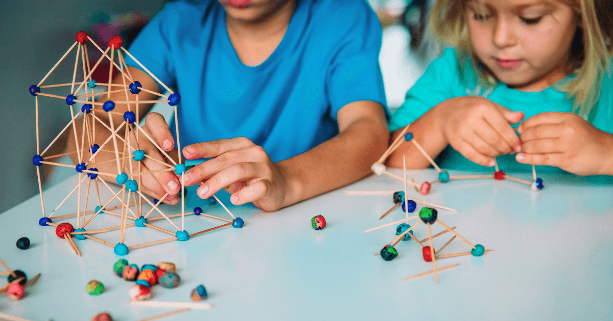 Two children build STEM structures on a table using toothpicks and colorful clay balls. The focus is on their hands as they assemble geometric shapes, fully engaged and concentrated on the creative activity.