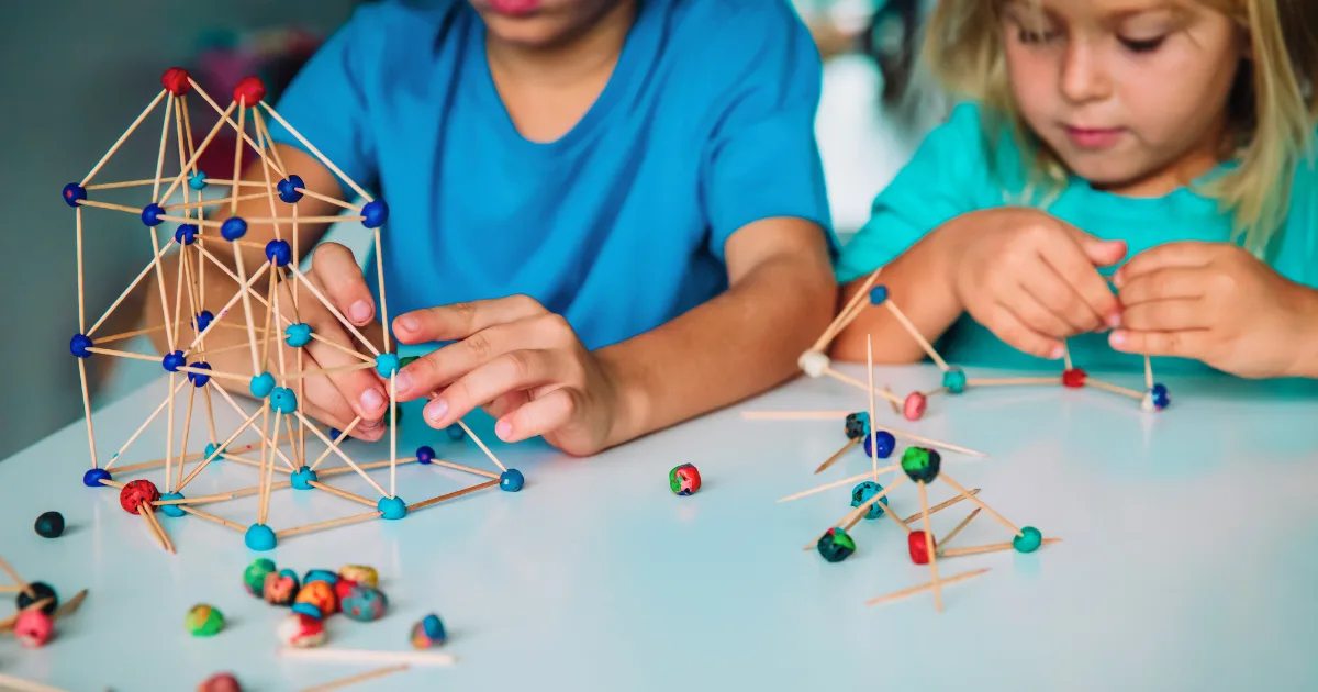 Two children build STEM structures on a table using toothpicks and colorful clay balls. The focus is on their hands as they assemble geometric shapes, fully engaged and concentrated on the creative activity.