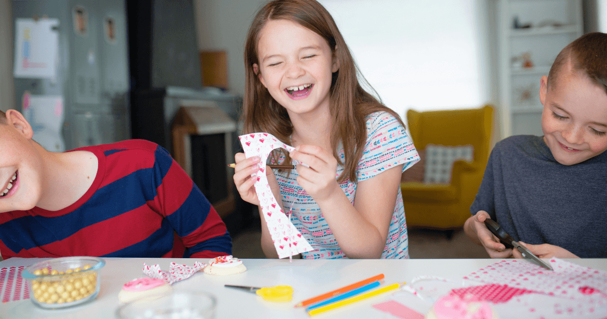 Three smiling children sit at a table doing Valentine's Day arts and crafts with patterned paper, scissors, and markers. The girl in the middle laughs while holding paper, and the boys beside her are focused on their crafts.