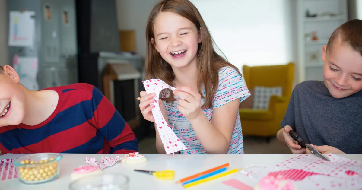 Three smiling children sit at a table doing Valentine's Day arts and crafts with patterned paper, scissors, and markers. The girl in the middle laughs while holding paper, and the boys beside her are focused on their crafts.