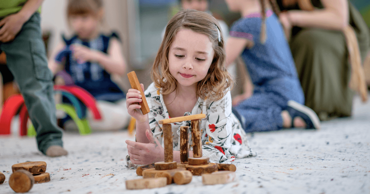 A young girl lies on the floor, concentrating as she builds a structure with wooden blocks—a hallmark of Waldorf education. Other children play in the blurred background, creating a lively indoor play area.