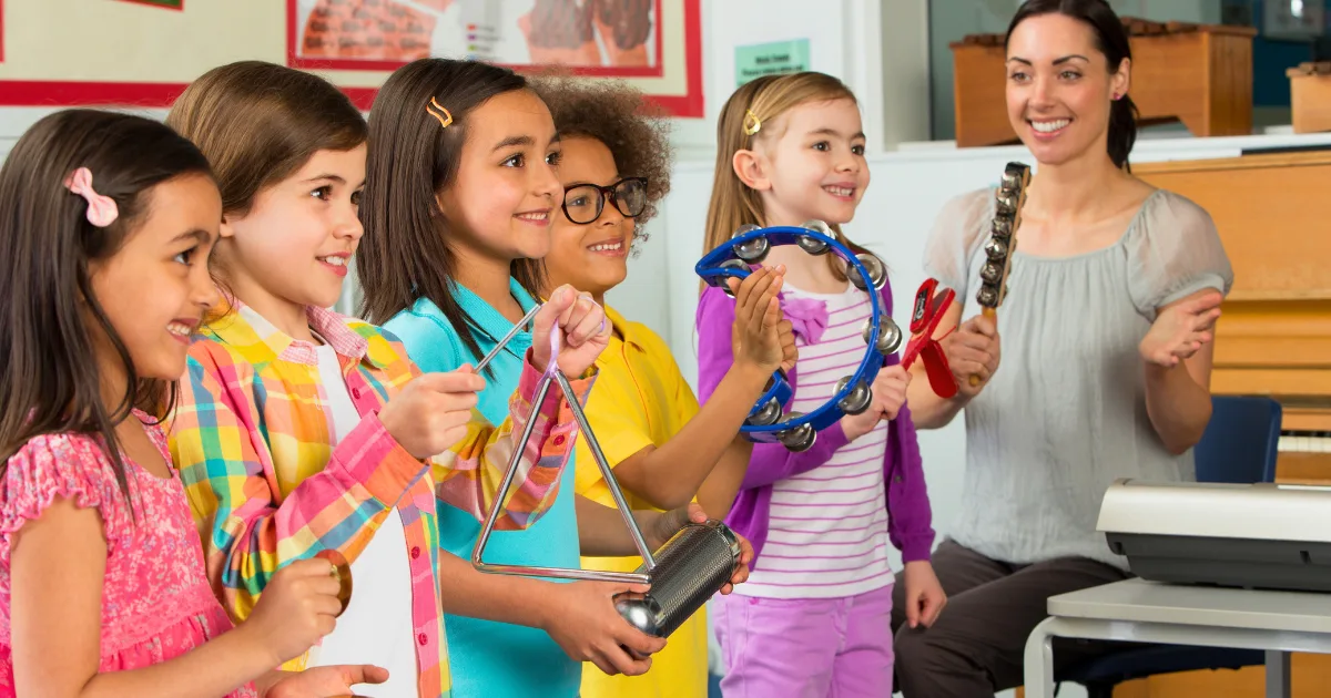 A group of smiling children stand in an affordable preschool classroom holding musical instruments, such as a triangle, tambourine, and maracas, while an adult woman, likely a teacher, sits nearby smiling and clapping.