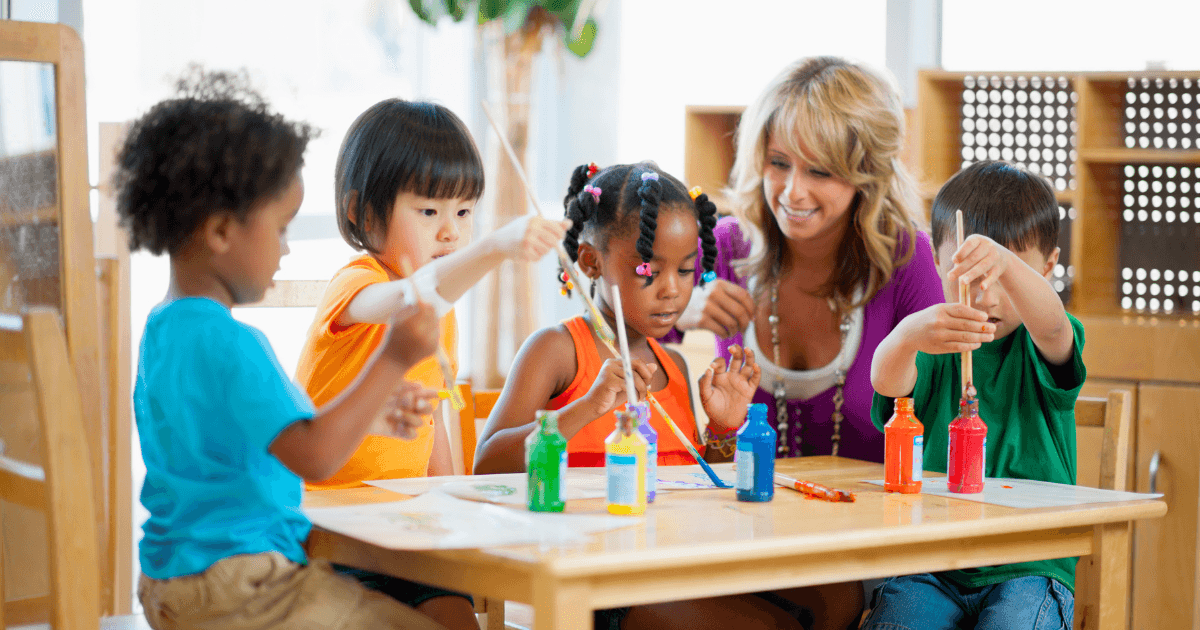 Four young children sit at a table painting with colorful paints and brushes, while a smiling adult woman watches and assists them in a bright, cheerful classroom that encourages authentic assessment.