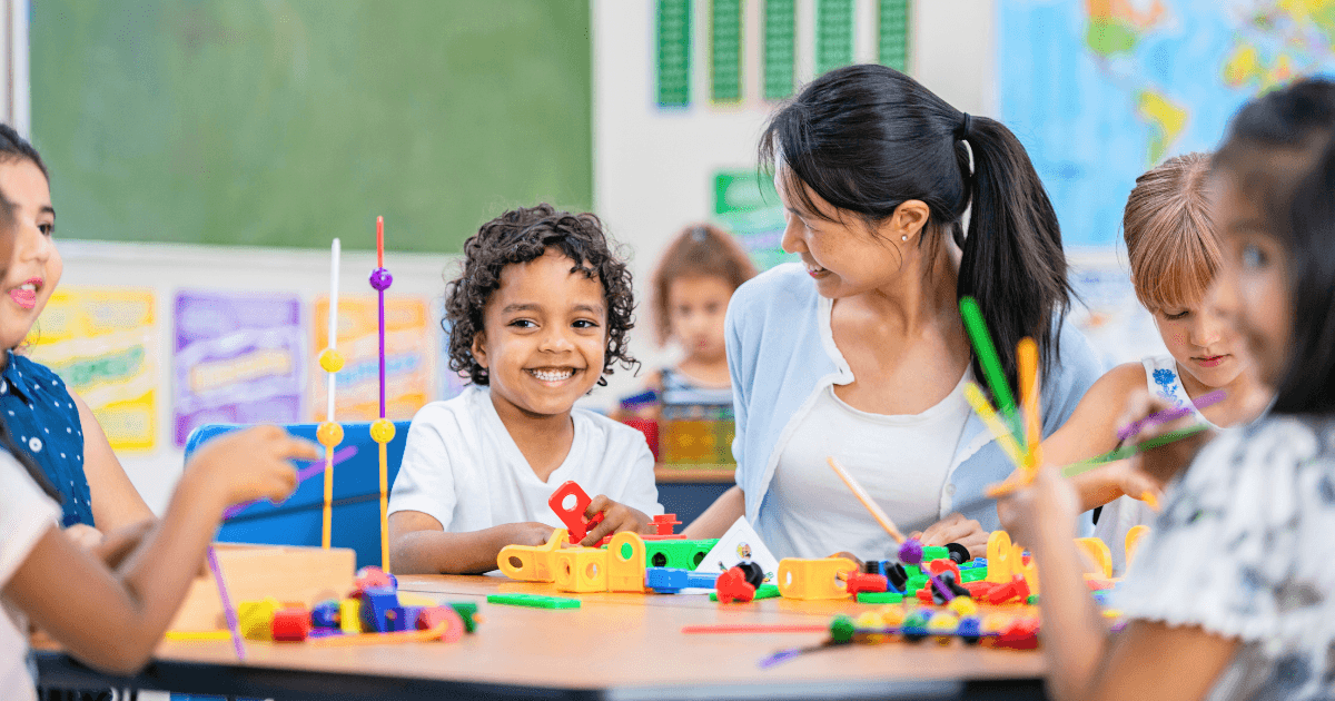 A group of young children and a female teacher sit around a classroom table, smiling and playing with colorful building toys, illustrating the benefits of early childhood education. Educational posters and a map are visible on the walls in the background.