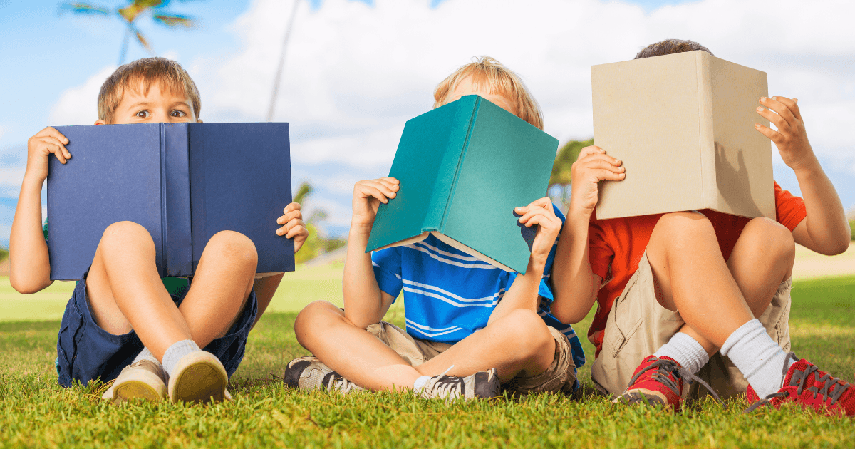 Three young children sit on grass outdoors, each holding books for early readers in front of their faces, hiding their expressions. The background features a bright sky and palm trees, suggesting a warm, sunny day.