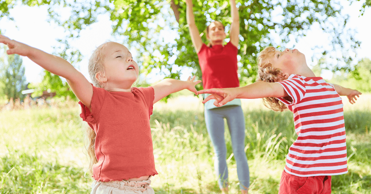 Two young children with arms outstretched stand outside in a sunny, grassy area, enjoying nature, while an adult in the background mimics their pose and guides them through simple breathing exercises under leafy green trees.
