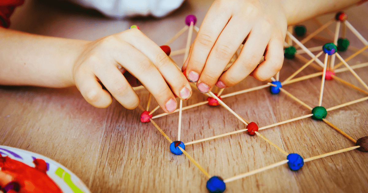 A child’s hands build a geometric structure using wooden sticks and colorful clay balls on a wooden table—a fun, budget-friendly STEM activity.