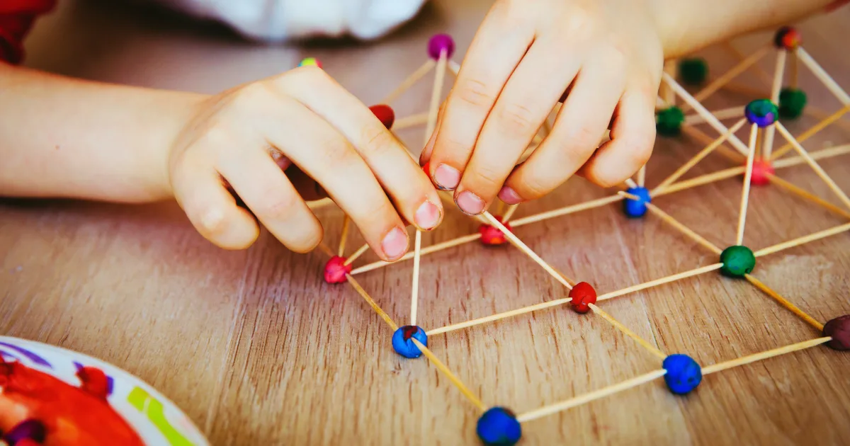 A child’s hands build a geometric structure using wooden sticks and colorful clay balls on a wooden table—a fun, budget-friendly STEM activity.