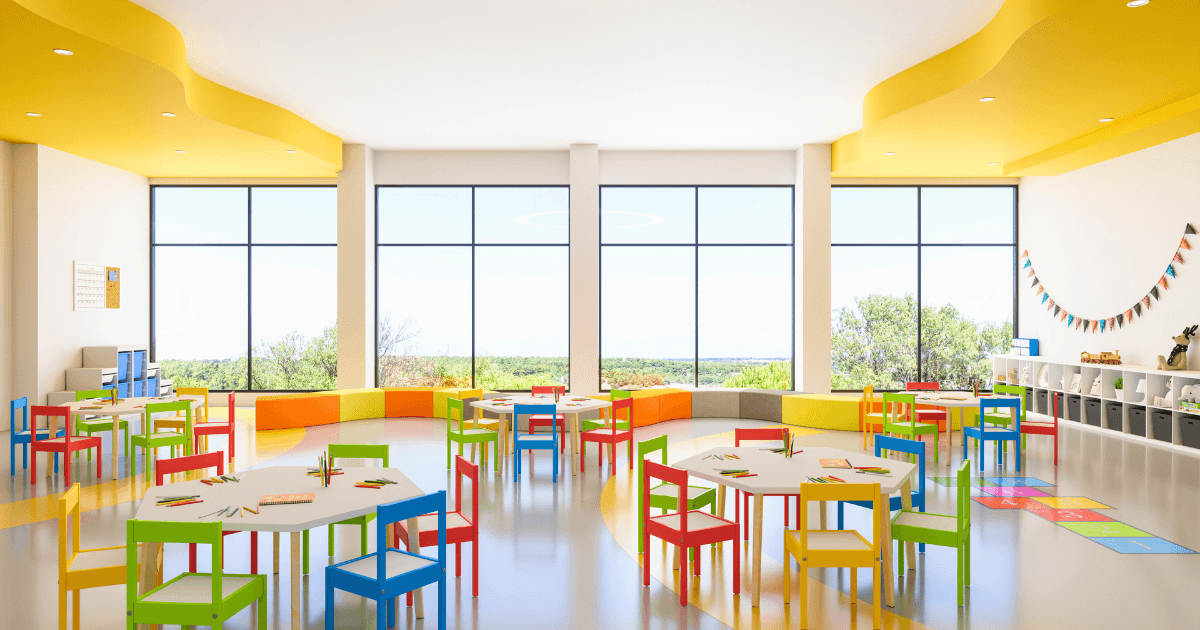 Bright, spacious classroom with large windows showcases thoughtful classroom design—colorful chairs and tables, various art supplies neatly arranged, playful wall decorations, and a view of greenery outside. The room is empty of people.