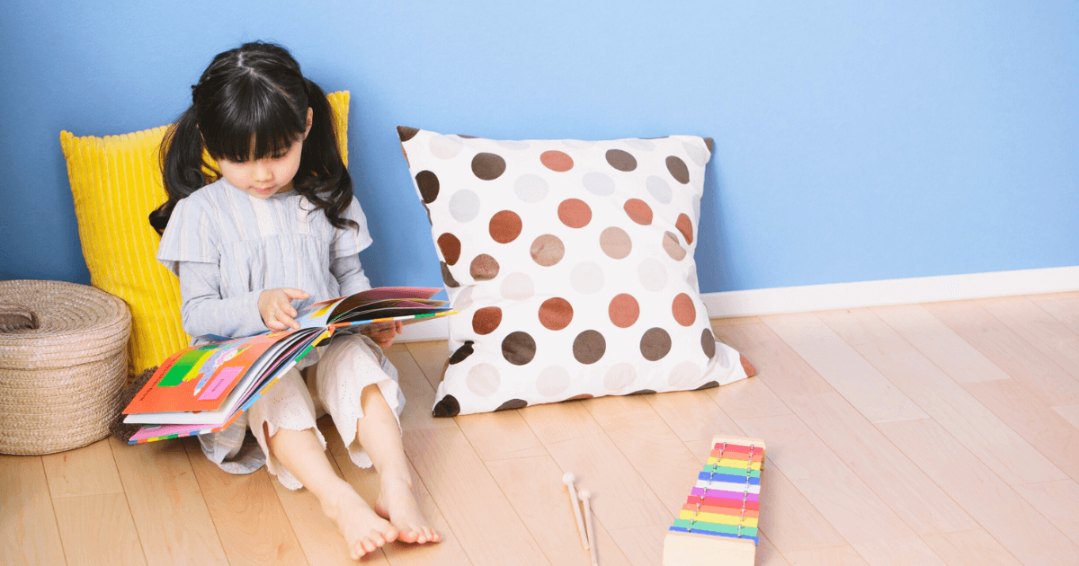 A young girl sits on the floor against pillows, reading a colorful book in her calm down corner. A xylophone lies nearby on the wooden floor, with a blue wall in the background creating a cozy and playful atmosphere.