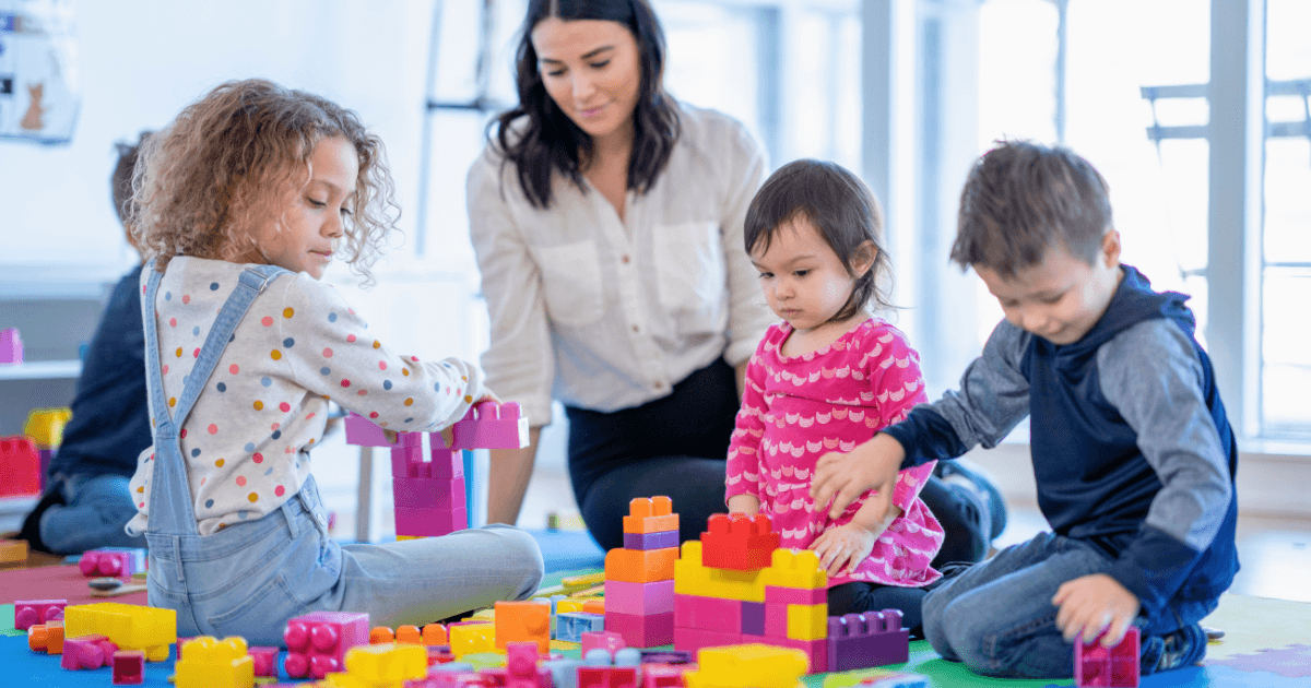 Three young children play with colorful building blocks on the floor while an adult woman watches and smiles, in a bright room with large windows—perfect for families seeking quality childcare near me.