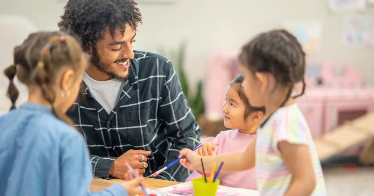 A man smiles while sitting at a table with three young girls who are painting and coloring together in a brightly lit room, highlighting one of the many positive childcare options compared to other settings. The atmosphere is cheerful and creative.