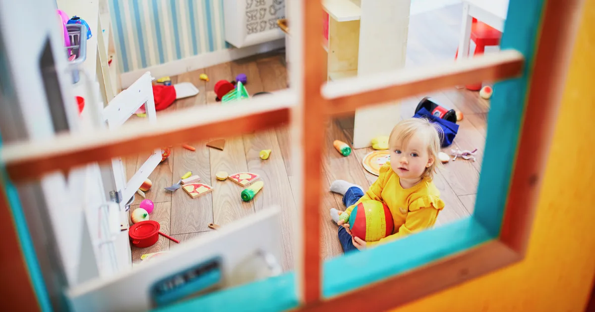 A toddler with blonde hair sits on the floor of a playroom holding a toy ball, surrounded by scattered toys and blocks, seen through a windowpane—a glimpse into engaging childcare options.