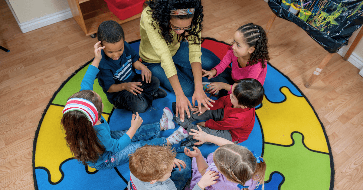 A teacher sits on a colorful puzzle-piece rug with six young children, leading a lively circle time activity. The children sit in a circle, reaching toward the teacher’s hands, and the atmosphere is interactive and joyful.