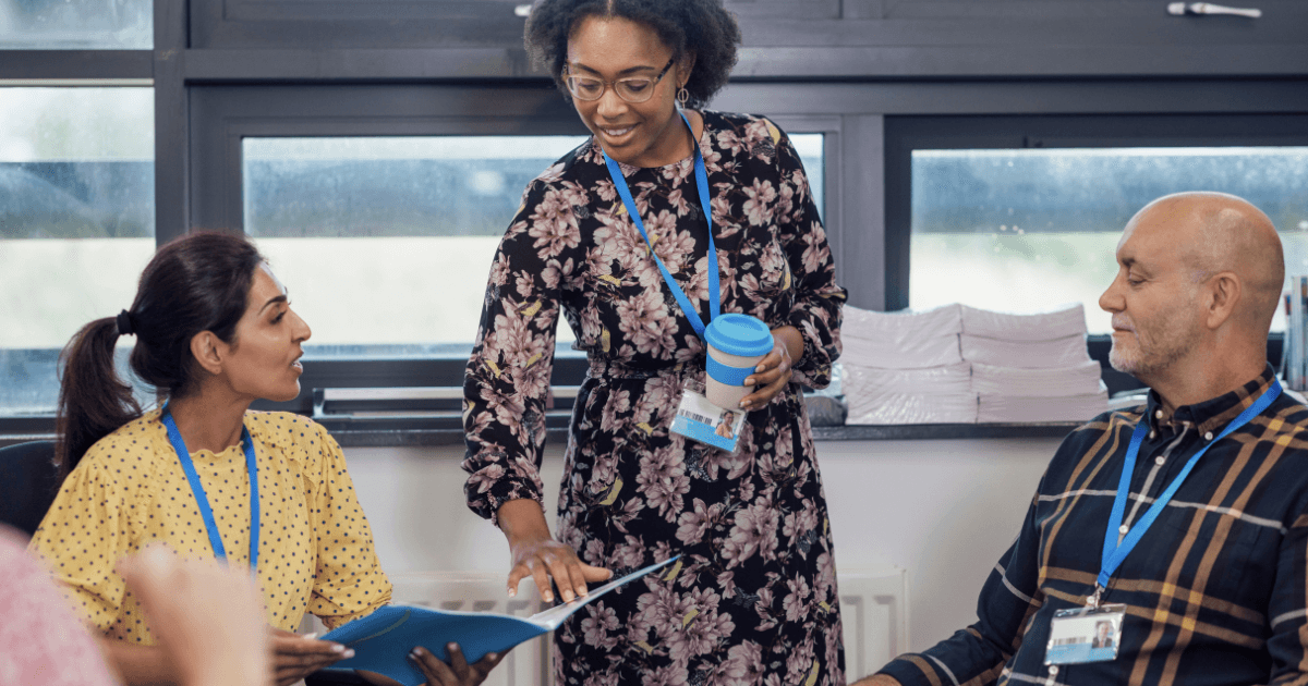 Three colleagues wearing ID badges are in discussion. A woman in a floral dress stands, holding a reusable cup and pointing to a document as she shares co-teaching strategies, while two seated people listen attentively amid stacks of paper in the office.
