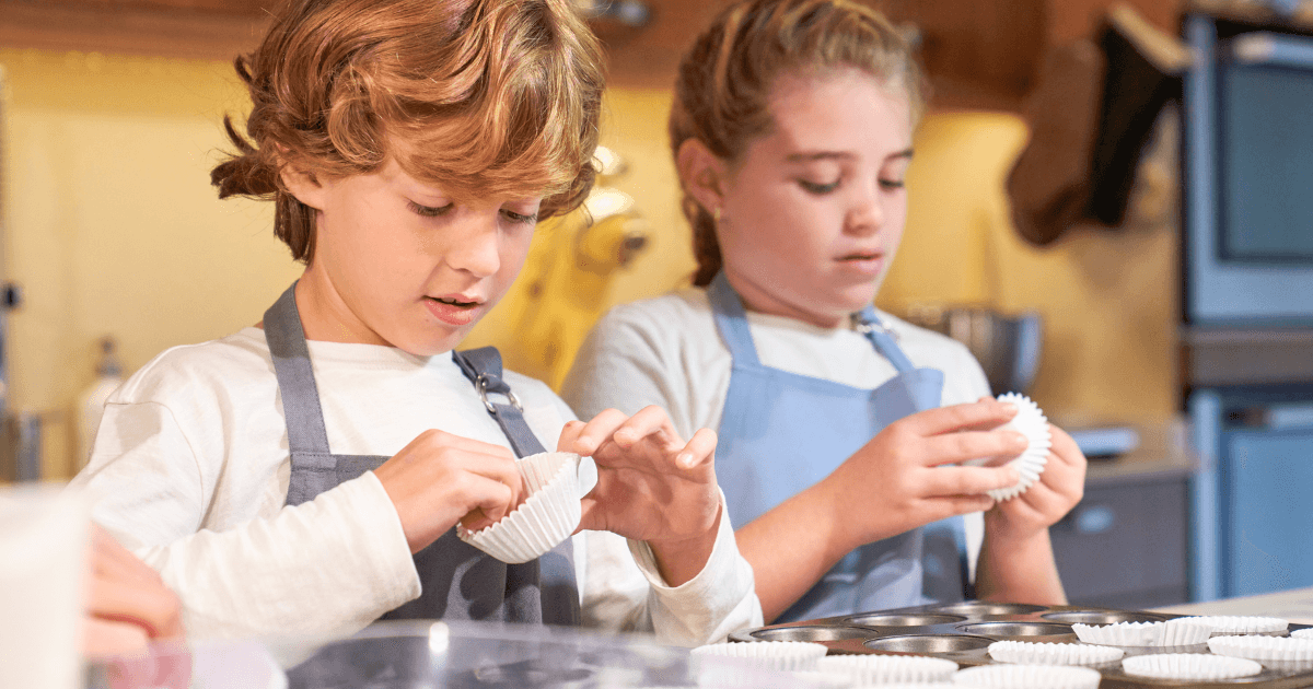 Two children in aprons, like little community helpers, prepare cupcake liners in a kitchen, carefully placing them into a muffin tray on the counter in front of them.