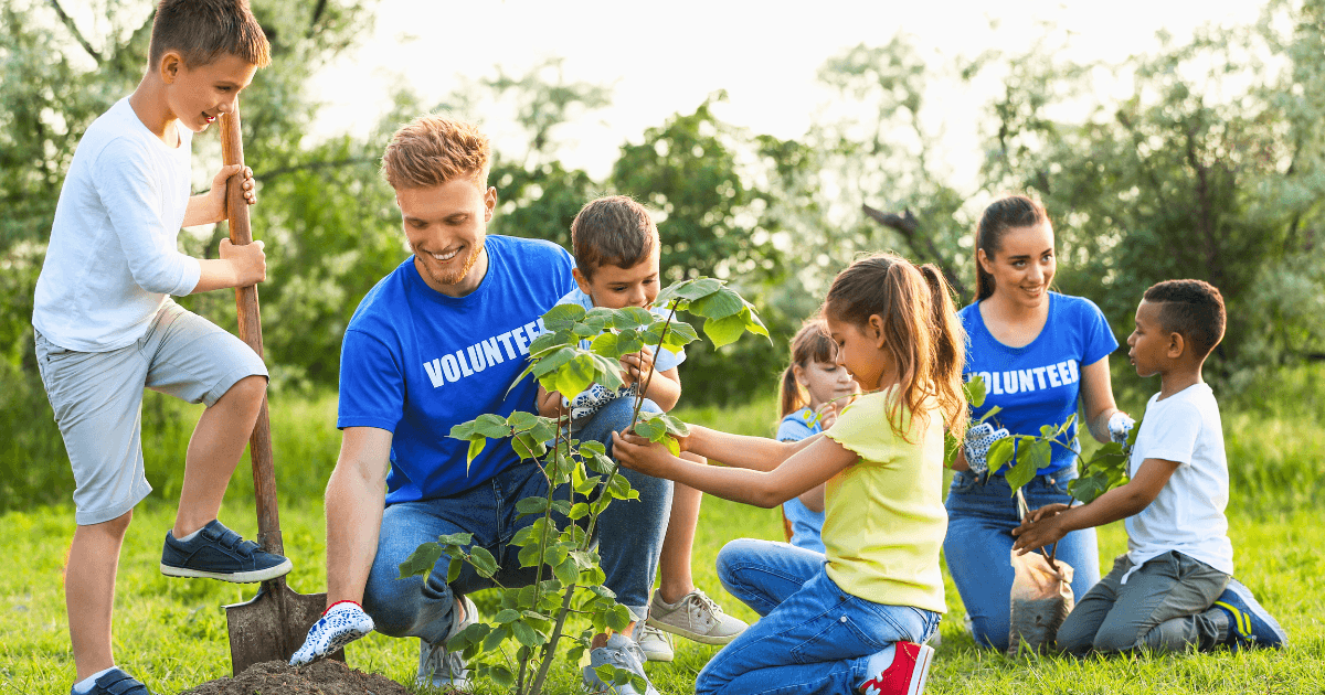 A group of community helpers and children work together outdoors, planting a tree. The adults and kids are smiling, wearing blue Volunteer shirts and gardening gloves, surrounded by green grass and trees.