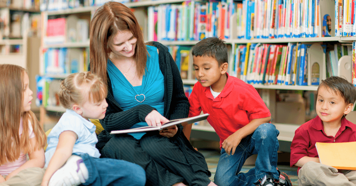 A woman sits on the floor in a library, reading a book to four young children gathered around her, listening attentively—a heartwarming scene that highlights the impact of community partnerships. Shelves filled with books are visible in the background.