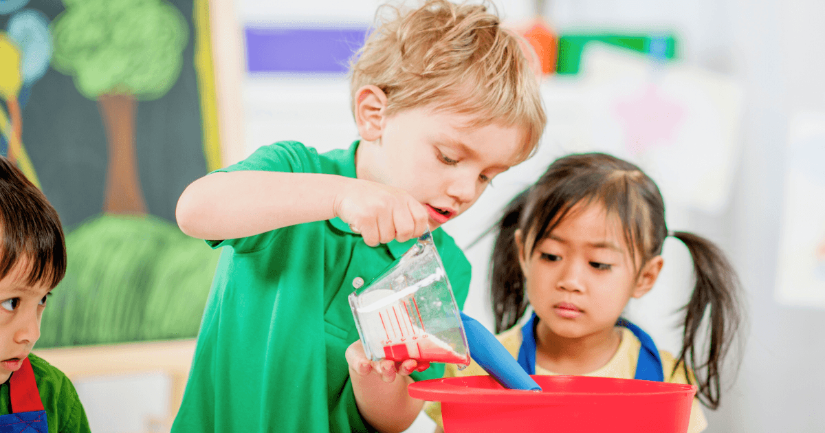 Three young children are engaged in an activity inspired by preschool curricula. One pours liquid from a measuring cup into a large red bowl while another watches, and a third child is partially visible on the left.