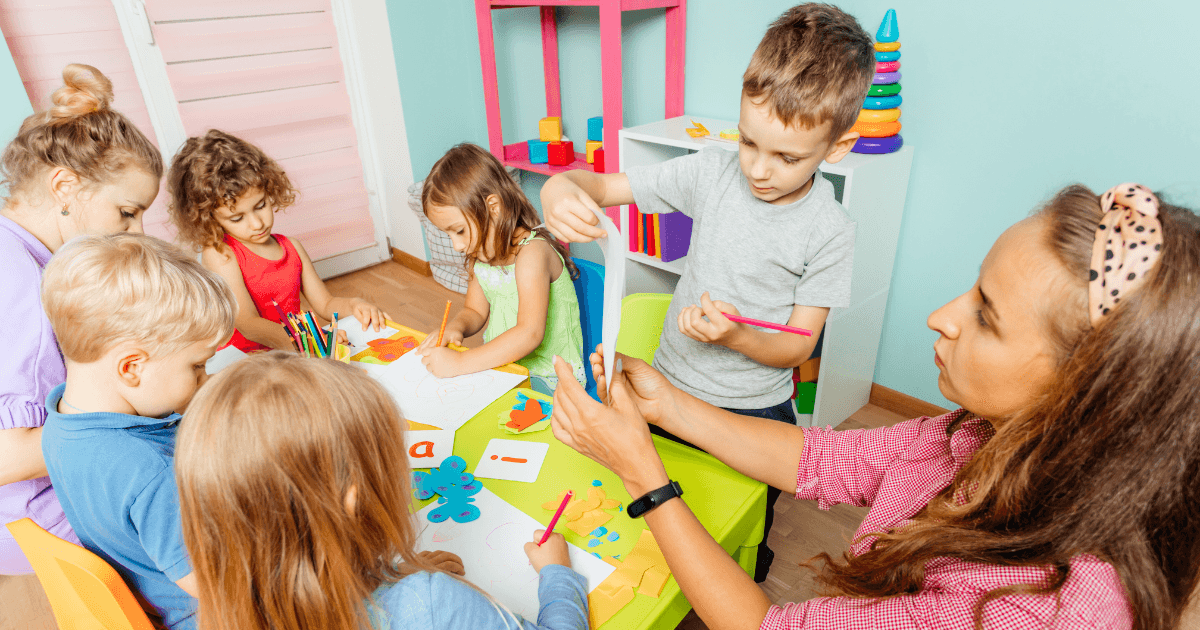 A teacher in a colorful classroom helps a group of young children with arts and crafts at a cooperative preschool. Children are drawing, using glue, and working with paper as the teacher assists one boy. Shelves and toys fill the background.
