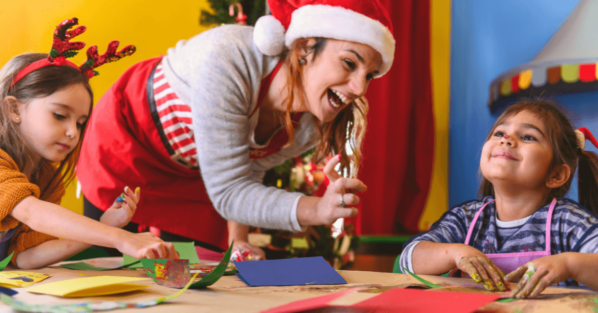 A smiling woman in a Santa hat helps two young girls with holiday crafts, sharing the joy of cultural celebrations at a table covered with colored paper and decorations. One girl wears reindeer antlers, while the other smiles up with paint on her hands.