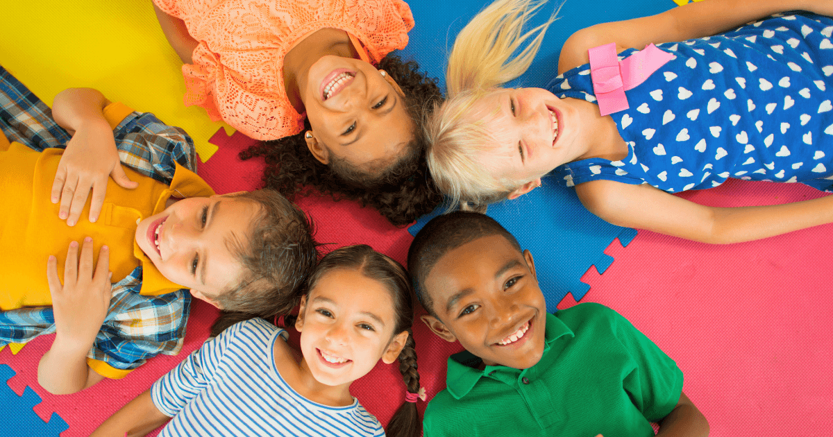 Five smiling children lie in a circle on colorful foam mats, showcasing cultural competence as they enjoy each other's company. Dressed in bright, casual clothes, they look happy and relaxed while gazing up at the camera.