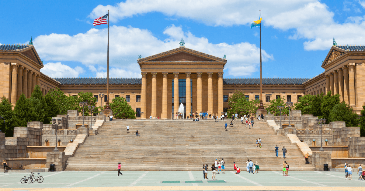 People walk up and down the wide stone steps leading to the Philadelphia Museum of Art on a sunny day, showcasing one of the city's premier cultural resources, with two large flagpoles and a historic building in the background.