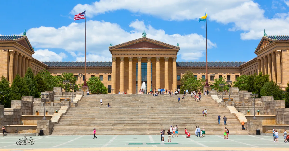 People walk up and down the wide stone steps leading to the Philadelphia Museum of Art on a sunny day, showcasing one of the city's premier cultural resources, with two large flagpoles and a historic building in the background.