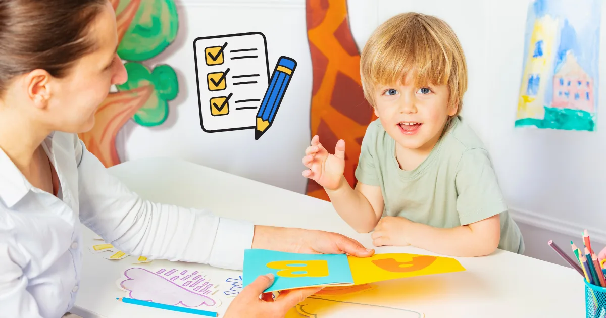 A young boy sits at a table with a woman, looking at colorful alphabet cards. The woman holds up a card with the letter A. Drawings and developmental checklists with a pencil are visible in the background.