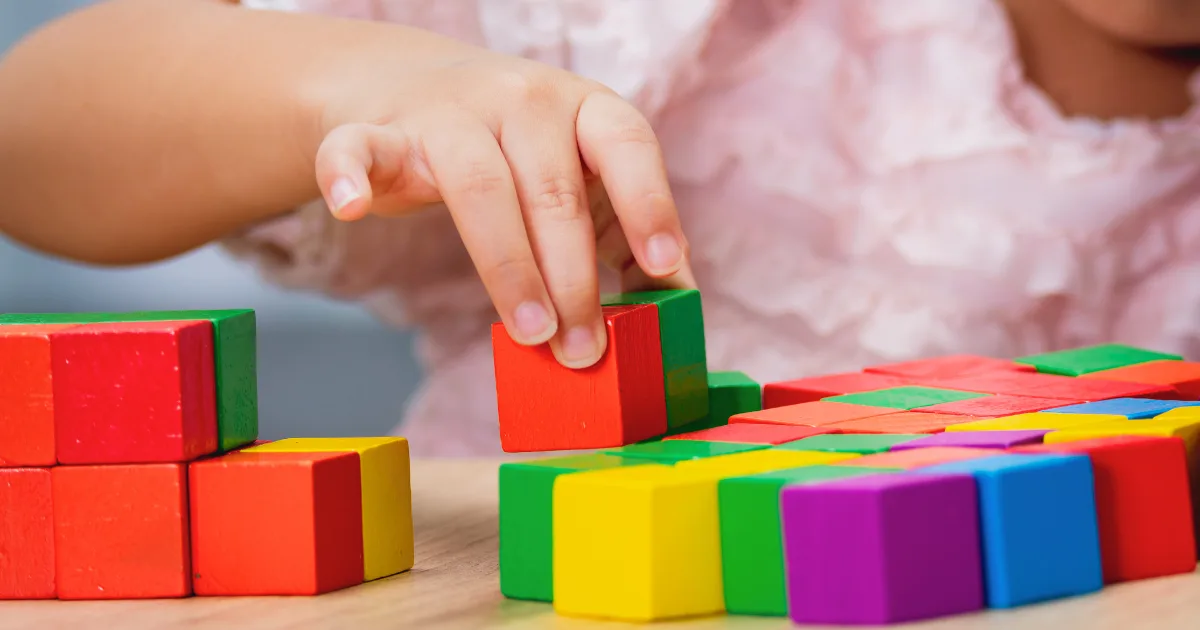 A child’s hand stacking colorful wooden blocks on a table, supporting key developmental milestones, with more blocks and a blurred pink shirt visible in the background.