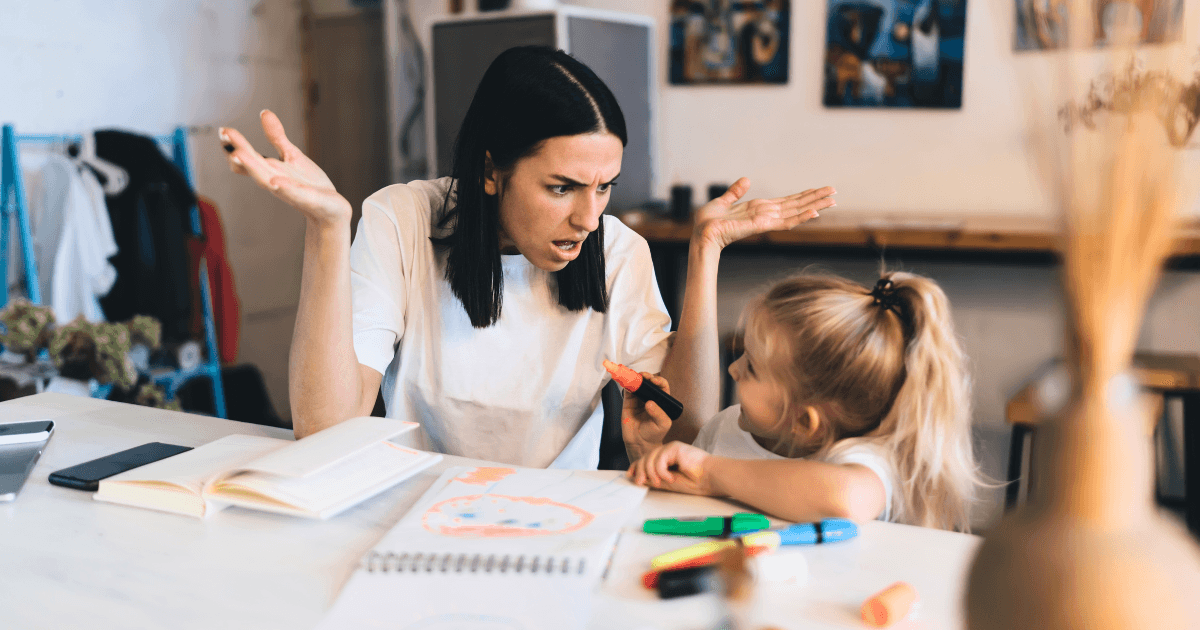 A frustrated woman with dark hair gestures with her hands while discussing developmental red flags with a young blonde girl sitting at a table covered with markers, paper, and drawings in a brightly lit room.