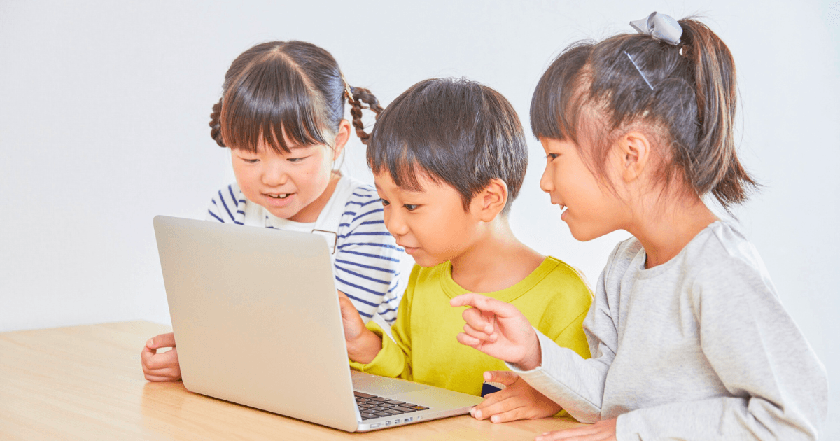 Three young children sit together at a table, smiling and looking at a laptop screen, appearing engaged as they explore the basics of digital citizenship.