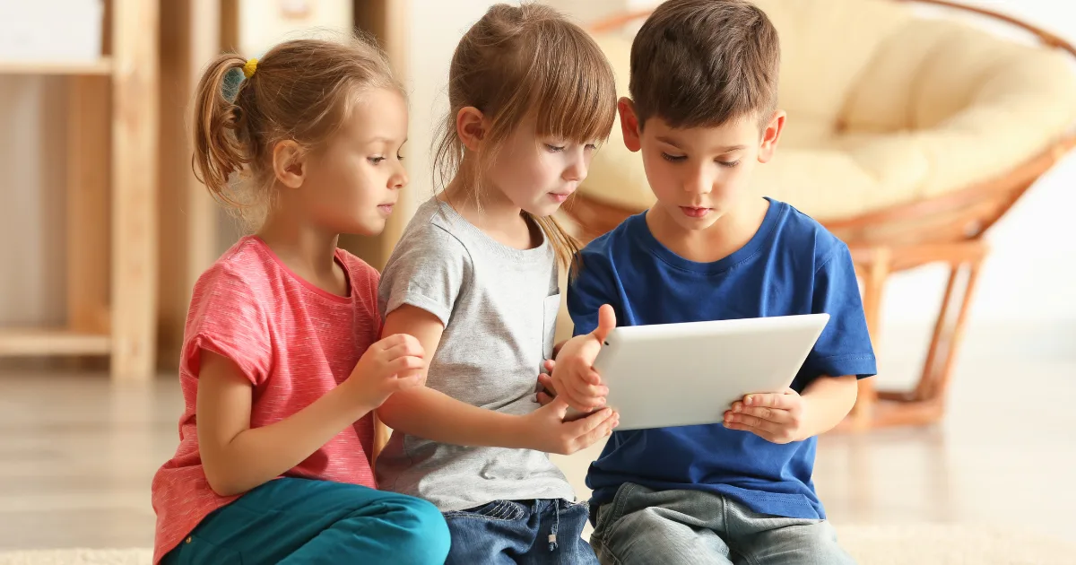 Three young children sit close together on the floor, looking at a tablet that the boy on the right is holding, appearing focused and engaged in digital storytelling. They are indoors in a cozy, softly lit room.