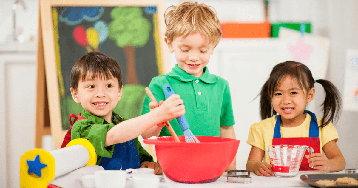 Three young children wearing aprons mix ingredients in a red bowl at a table, smiling. A chalkboard with colorful drawings is in the background, enhancing the playful, classroom atmosphere with dramatic play themes.