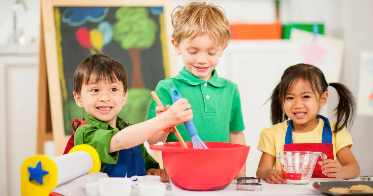 Three young children wearing aprons mix ingredients in a red bowl at a table, smiling. A chalkboard with colorful drawings is in the background, enhancing the playful, classroom atmosphere with dramatic play themes.